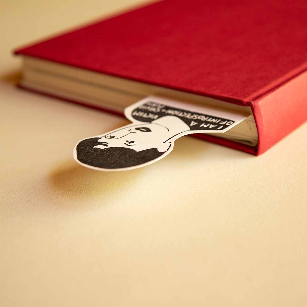 Un libro de tapa dura de color rojo con un marcapáginas de dibujos animados en el que aparece un hombre con bigote asomándose, sobre un fondo beige.
