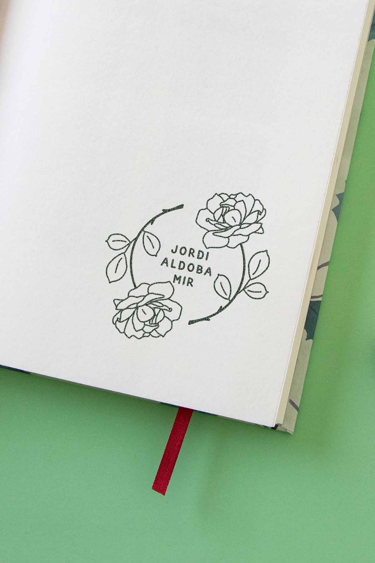 Un sencillo boceto de un diseño floral con rosas con los nombres “jordba” y “aldmir” dentro de un círculo, dibujado en un cuaderno abierto con un marcapáginas rojo, colocado sobre un Ex-libris Corona de rosas verde de Les Tampons de Roser.