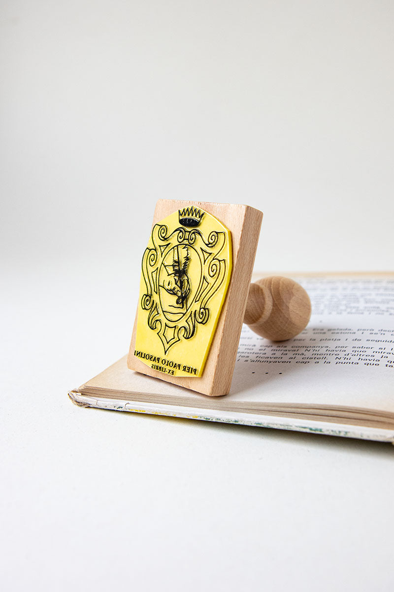 Sello ex-libris con un escudo heráldico con una corona, dentro del escudo se ve una mano escribiendo con una pluma