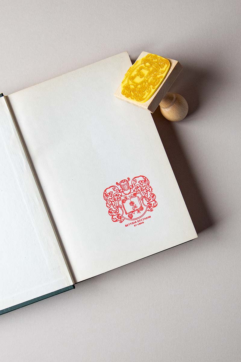 Portadilla de libro estampada con un escudo heráldico con yelmo en el interior hay un libro del que salen rosas