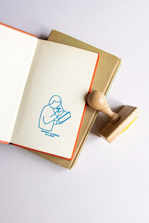 Portadilla de libro estampada con una lectora leyendo y oliendo una rosa