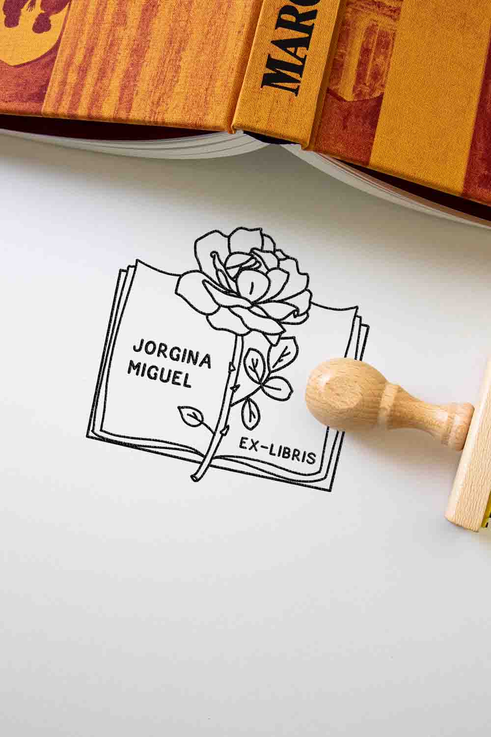 Un Ex-libris Libro abierto con rosa personalizado de Les Tampons de Roser que presenta un diseño de rosa con los nombres "Jorgina Miguel" impresos, sobre una superficie blanca junto a un sello de goma de madera y una pila de libros.