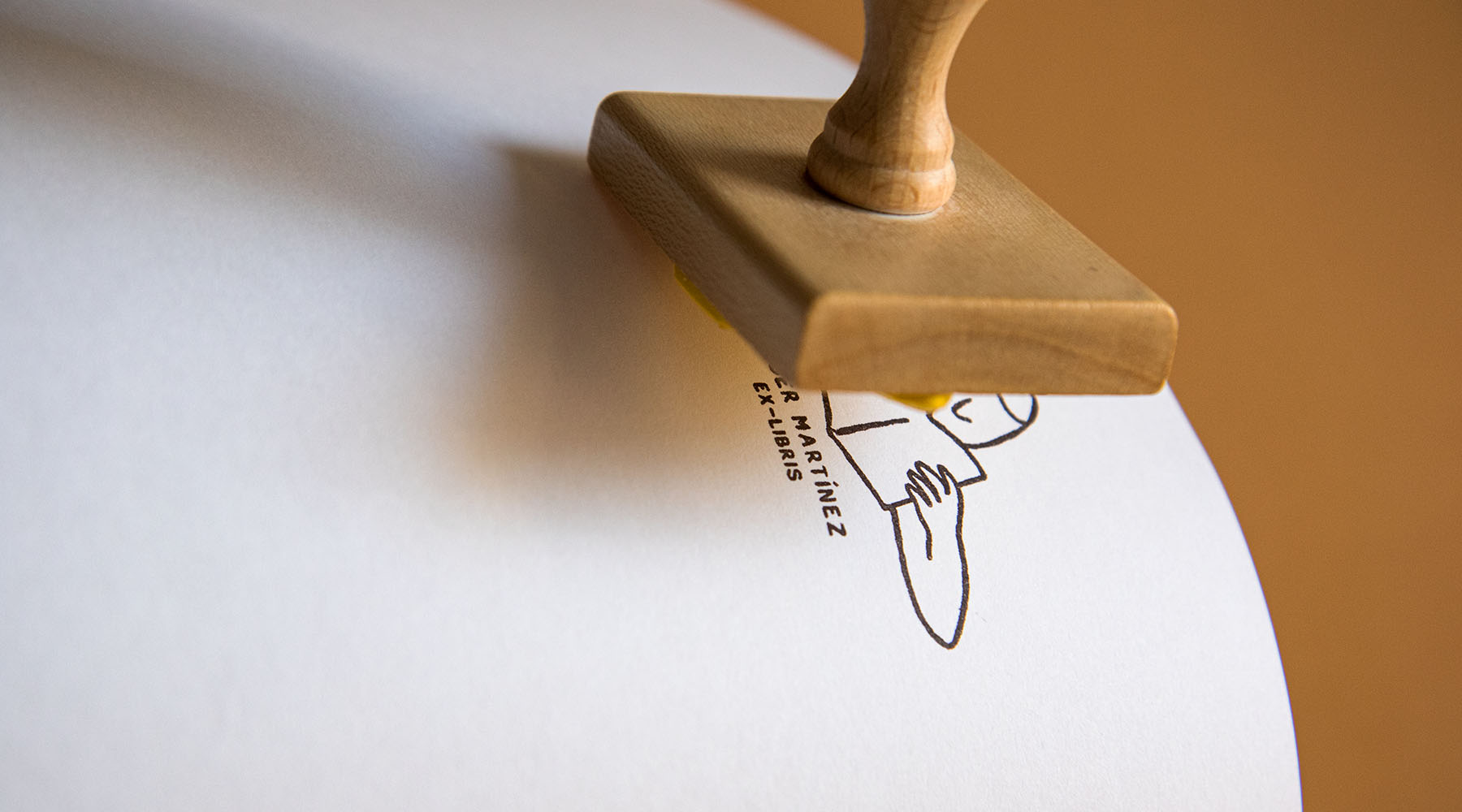 Un sello de madera prensado sobre papel, que deja una impresión en tinta de un dibujo lineal minimalista de una cara con el texto "a. martinez.