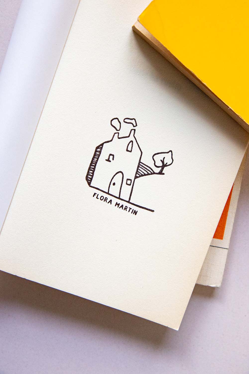 Portadilla de libro estampada con una casa en el campo con humo saliendo de la chimenea y un árbol al lado