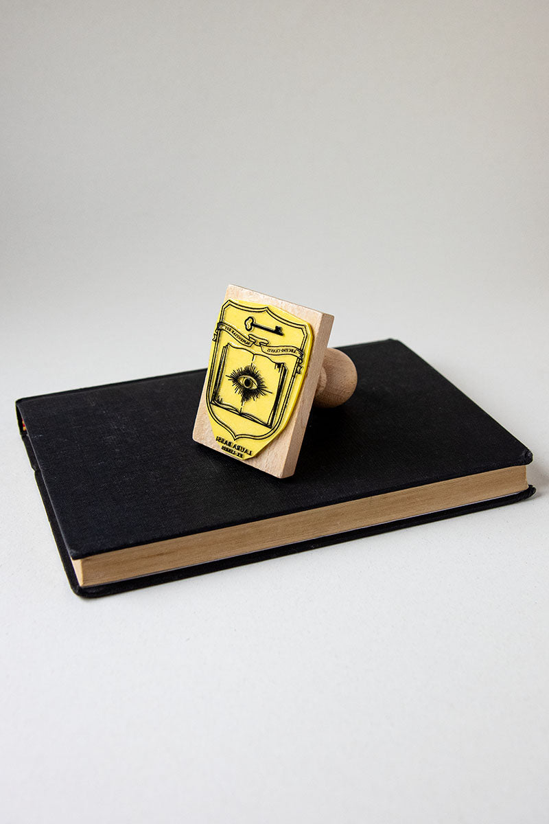Sello ex-libris con un escudo heráldico donde aparece un libro abierto con un ojo enmedio y una llave, enmedio hay una banderola donde aparece la cita "lectio clavis sapientiae est"