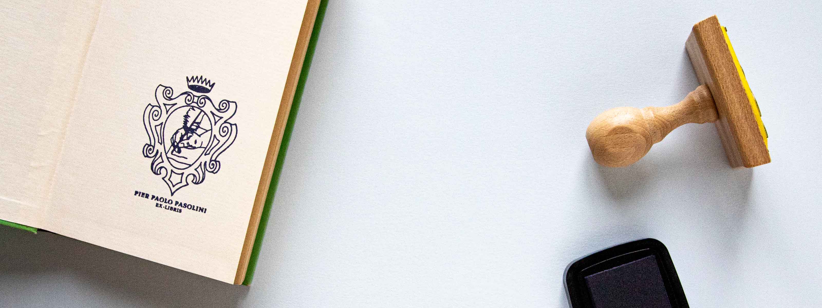 Exlibris personalizado sellado en la página de portada de un libro abierto con un escudo heráldico con corona, dentro del escudo se ve una mano escribiendo con una pluma
