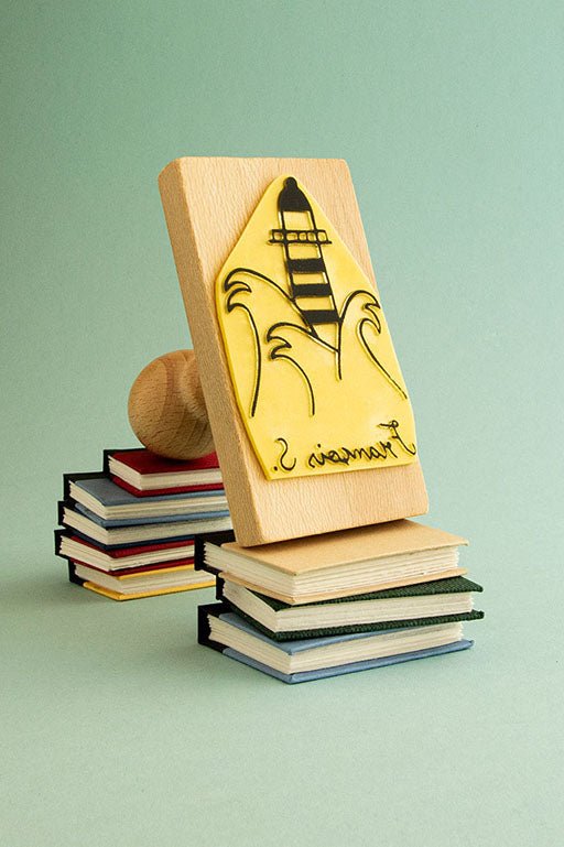 Un sello de madera de Les Tampons de Roser con un diseño de cohete, que presenta el libro Ex-libris Al faro de Virginia Woolf, apoyado sobre una pequeña pila de libros coloridos sobre un fondo verde claro.
