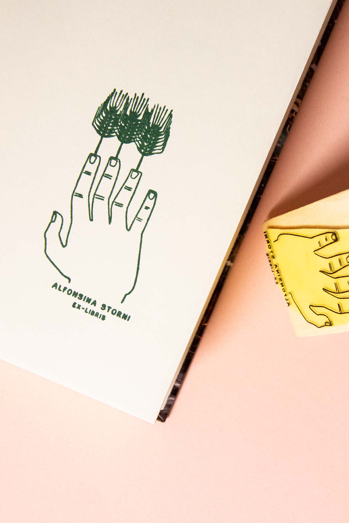Una ilustración dibujada a mano de una mano con árboles como dedos sobre papel blanco, junto a un sello de madera amarillo sobre un fondo melocotón. El texto "Les Tampons de Roser Ex-libris Dedos de trigo