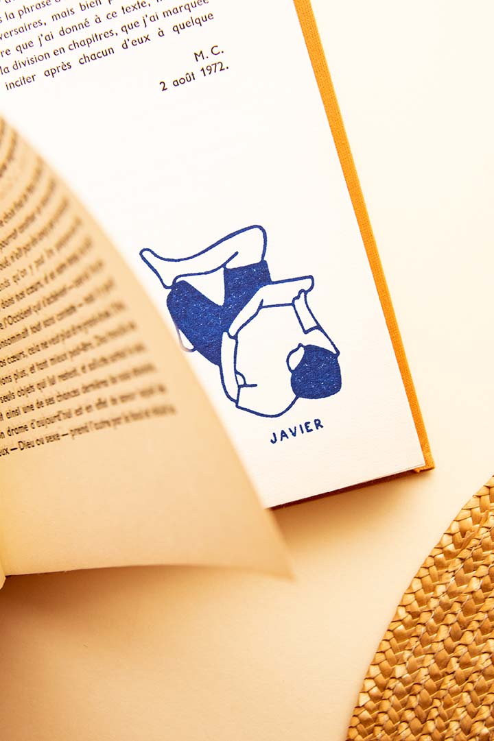 Un libro abierto con un marcador azul que presenta un dibujo estilizado de una línea blanca de un lector leyendo, con la etiqueta "Ex-libris Lector". Se ven parcialmente los bordes de la página y un sombrero de paja, de Les Tampons de Roser.