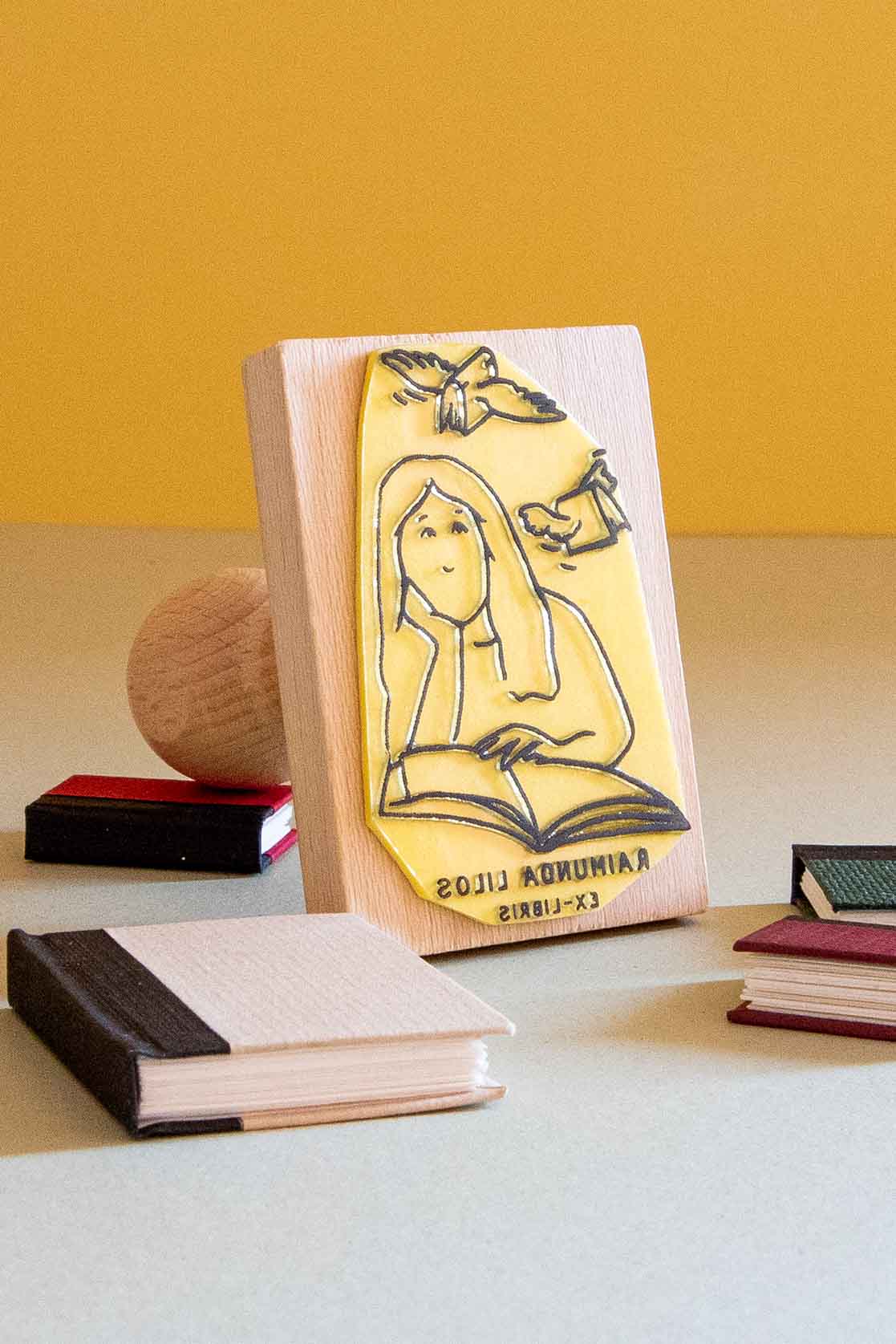 Sello exlibris grabado con una mujer leyendo un libro, con dos libros volando sobre ella