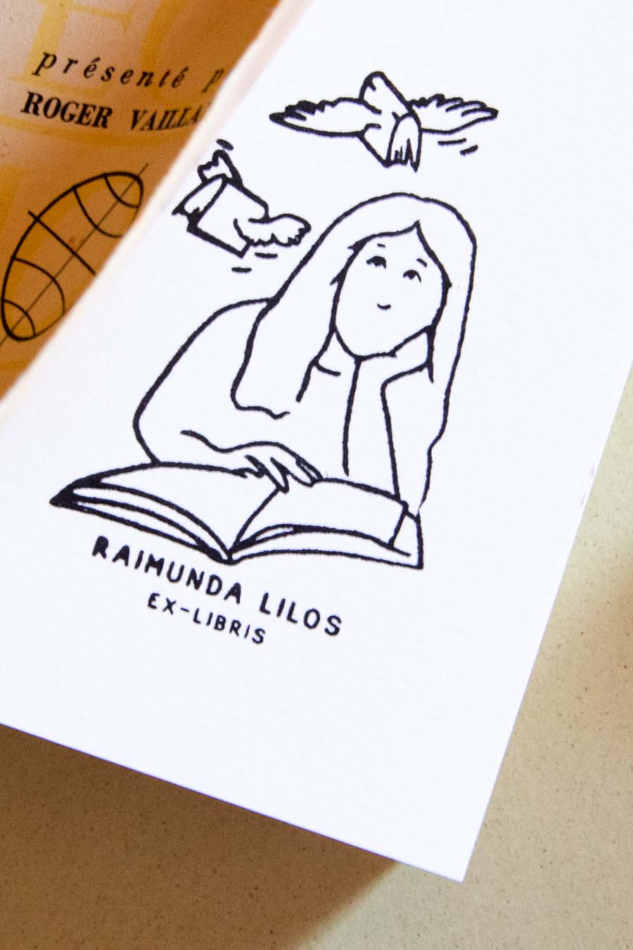 Portadilla de libro estampada con sello exlibris de una mujer leyendo un libro, con dos libros volando sobre ella