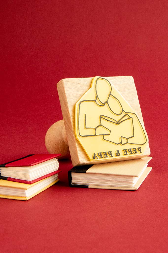 Frase con producto reemplazado: Un Ex-libris Los amantes geométricos con el ícono de una persona leyendo un libro y el texto "read & learn" se alza sobre un fondo rojo, al lado de una pequeña pila de libros en miniatura. Este Les Tampones de Roser