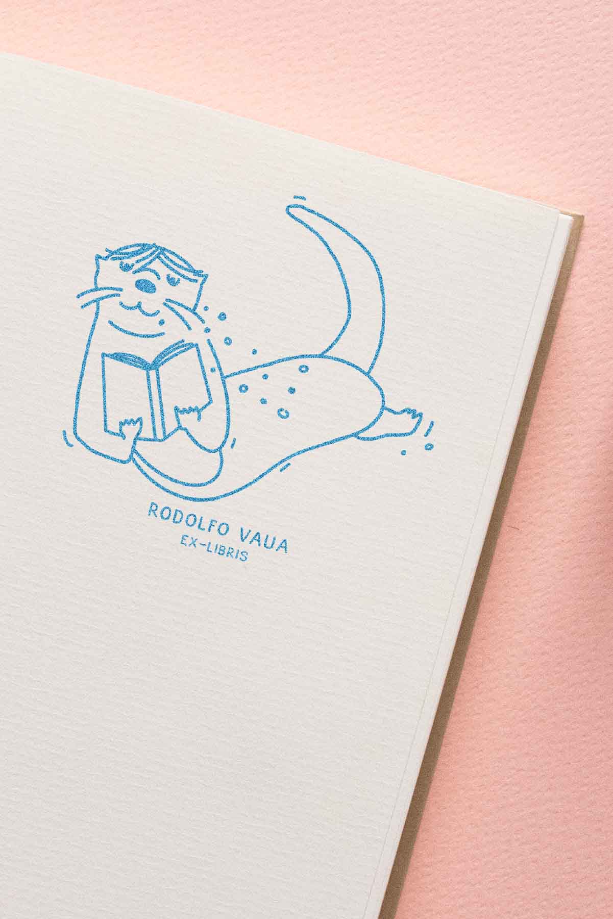 Ilustración de un gato contento tumbado boca arriba leyendo un libro, con "Les Tampons de Roser Ex-libris Nutria lectora" escrito debajo, sobre un papel texturizado sobre un fondo rosa.