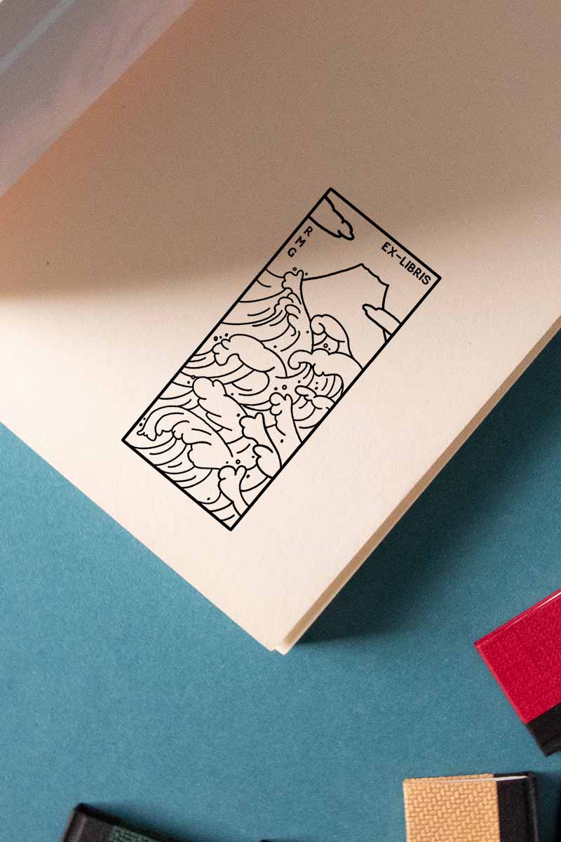 Un Ex-libris Olas con una detallada ilustración en blanco y negro de la Gran Ola de Kanagawa se coloca dentro de un libro abierto con un fondo de papel de colores de Les Tampons de Roser.