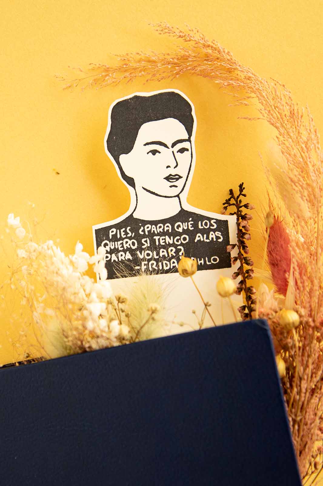 A Marcapáginas Frida de Les Tampons de Roser sobre un fondo amarillo, con una cita en español y rodeada de flores secas, colocada encima de un libro azul.
