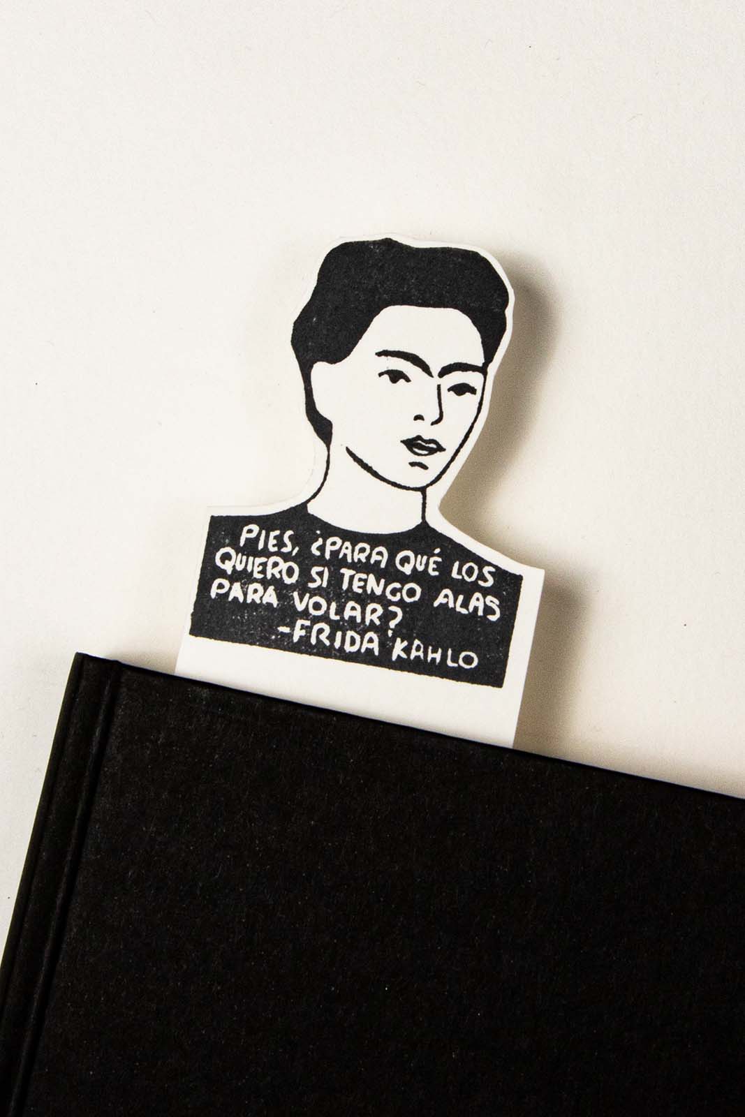 A Marcapáginas Frida de Les Tampons de Roser con su retrato y una cita en español, apoyada en el borde de un cuaderno negro sobre un fondo blanco.