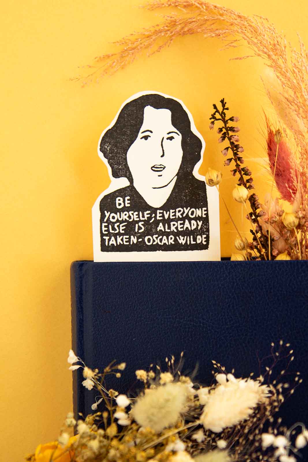 Un montaje decorativo que presenta un Marcapáginas Oscar Wilde de Les Tampons de Roser que muestra el retrato y la cita literaria de Oscar Wilde, rodeado de flores silvestres sobre un fondo amarillo.